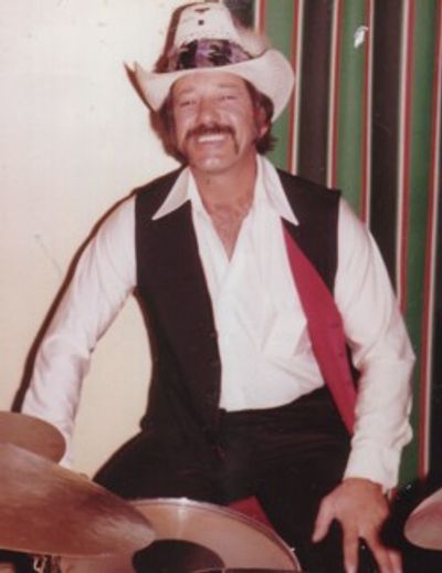 Man in cowboy hat and vest smiling while seated behind drums.
