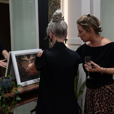 Two women examining a framed artwork indoors, one holding a glass of wine.