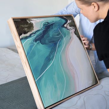 Person holding a framed abstract painting of a river or coastline.