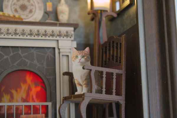 A cat figurine sitting on a miniature chair by a fireplace.