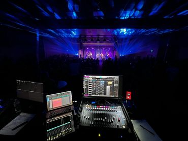 Sound mixing console and screens at a live concert with blue stage lighting.