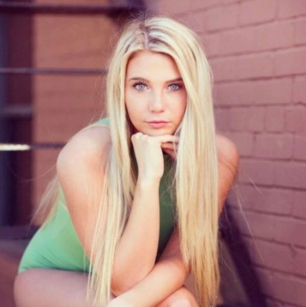 Young woman with long blonde hair sitting against a brick wall.