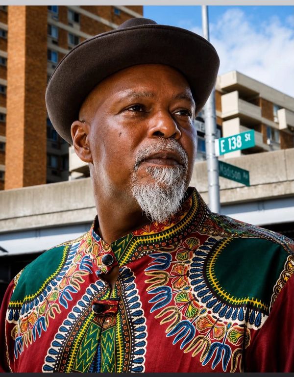 Man with a gray beard wearing a colorful patterned shirt and brown hat on city street.