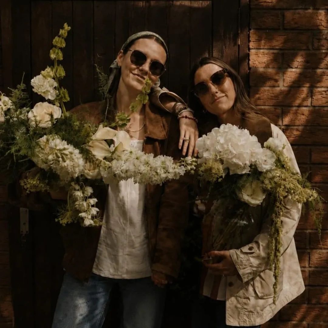 Two women holding flower bouquets, smiling with sunglasses.