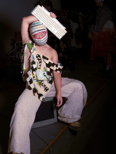 Person in colorful knitwear mask holding a keyboard, sitting on an old monitor.