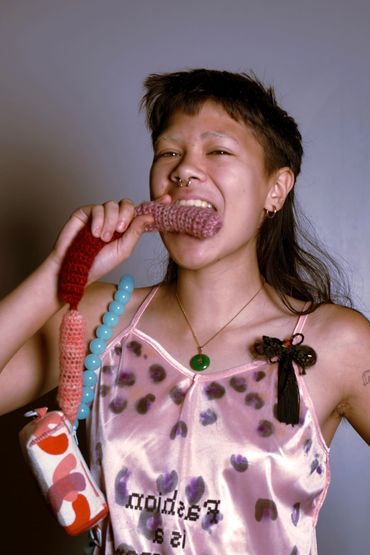 Person playfully biting a knitted item, wearing a satin top with text and accessories.