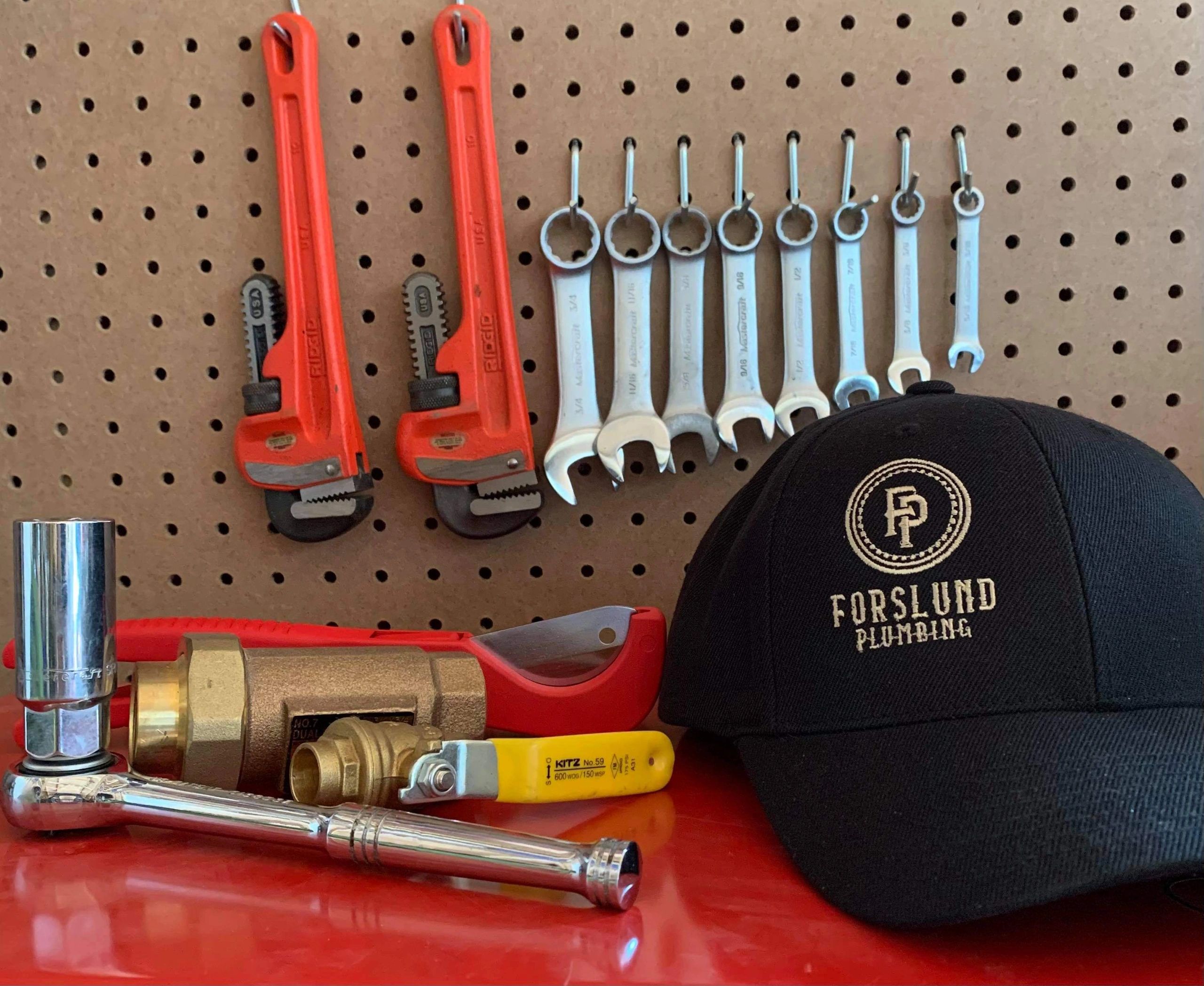 Forslund Plumbing logo black hat beside plumbing hardware on a red workbench