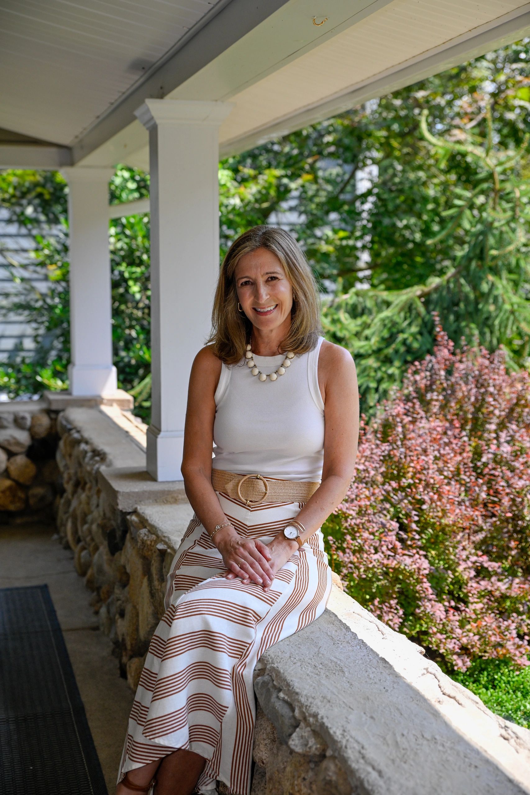 Smiling woman in white top and striped skirt sits on a porch with greenery in the background.