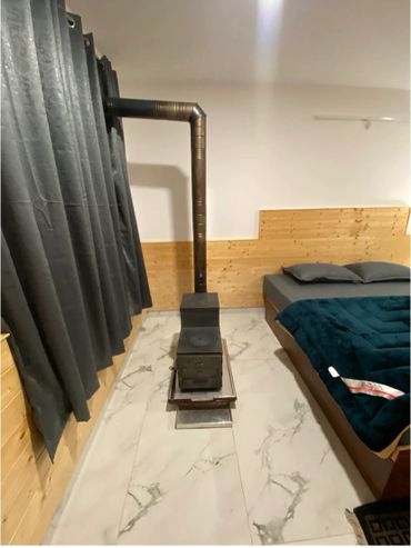 Compact wood stove in a cozy bedroom with wooden paneling and dark curtains.