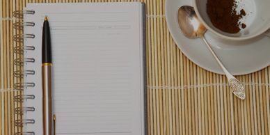 Notebook with pen beside a cup of coffee on a bamboo mat.