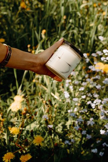 Bougie 8 oz Bruine océanique Émi Bougie. Main qui tient une bougie au-dessus des fleurs.