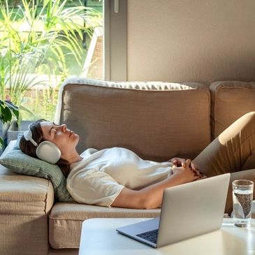Woman relaxing on a beige couch with headphones, laptop, and glass of water nearby.