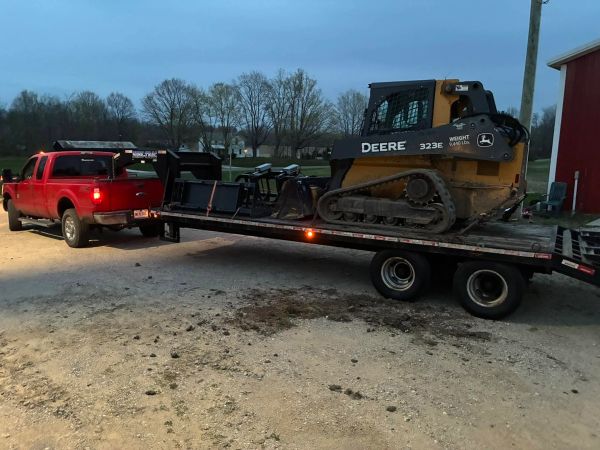 Red truck towing a flatbed trailer with a John Deere 323E skid steer.