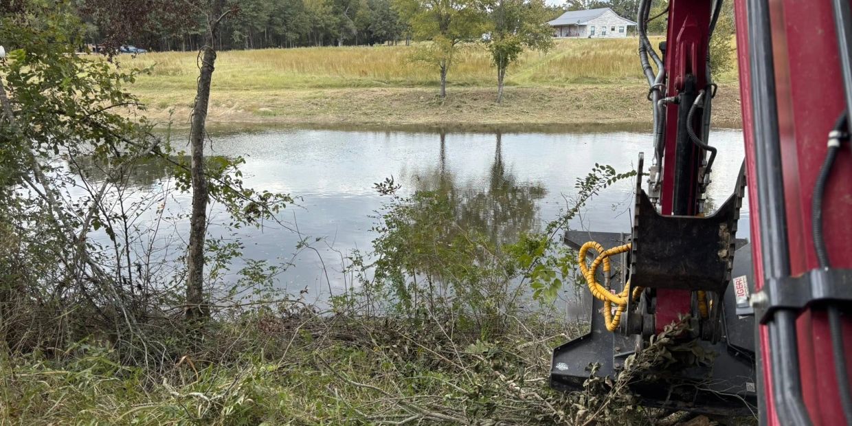 Excavator digging near a pond with grassy fields and trees.