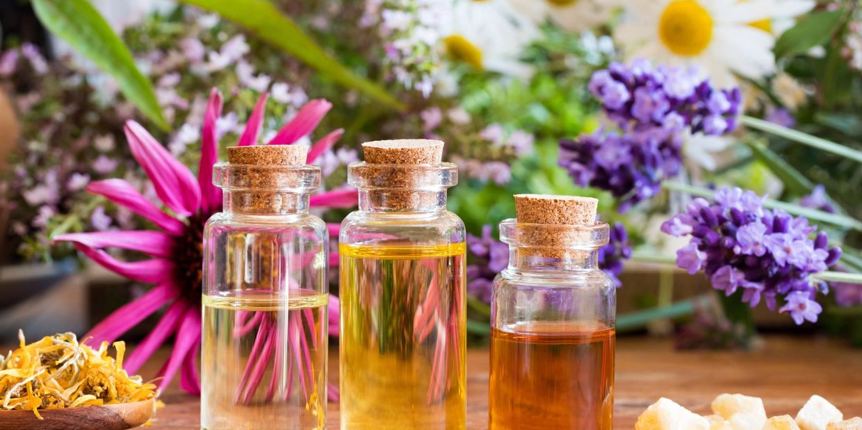 Three glass bottles with corks containing essential oils in front of colorful flowers.