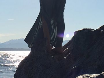 Person standing on a rock by the sea at sunset, wearing a flowing dress.