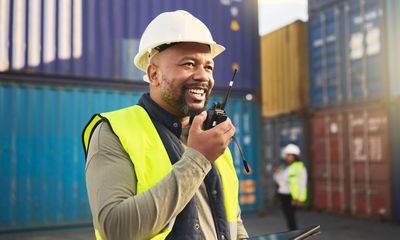 Smiling worker in safety gear using a walkie-talkie at a shipping yard.
