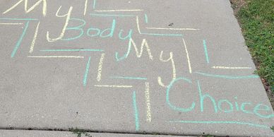 The words "My Body My Choice" written on a sidewalk in yellow and green chalk.