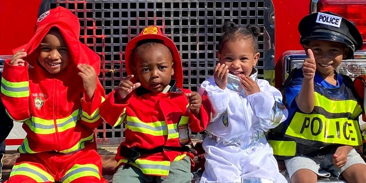 Four children sitting in front of a fire truck. They are dressed as firefighters and polic officers.