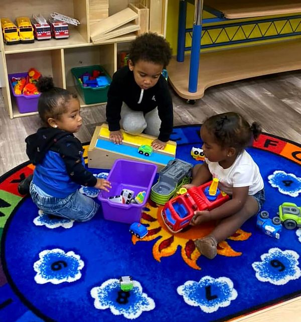 Three young students sit on a bright blue carpet while playing with toy trucks.