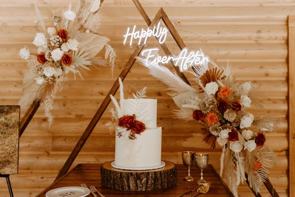 white wedding cake on a log stand in front of floral arrangements and a happily ever after sign