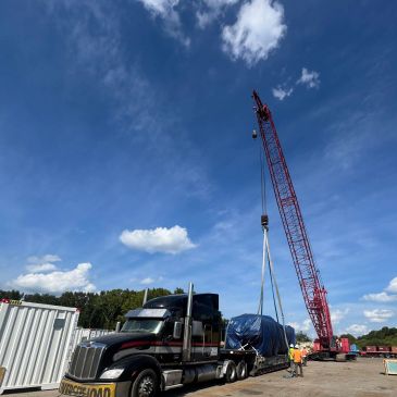A crane lifts a covered load onto a flatbed truck under a blue sky.
