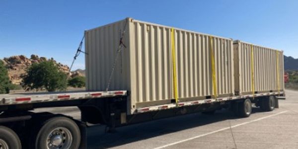 Flatbed trailer carrying two large beige shipping containers under a clear blue sky.