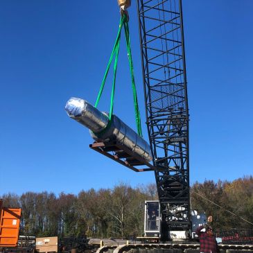 Crane lifting large cylindrical metal object with green straps outdoors.