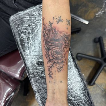 Forearm tattoo with books, flowers, fireflies, and a compass design.