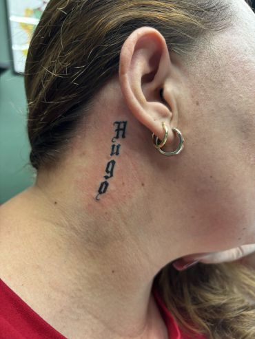 Tattoo of the word 'Hugo' behind the ear with earrings.