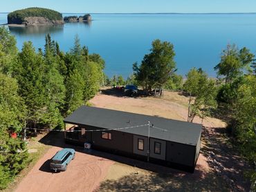 A modern cabin with a car parked nearby, surrounded by trees, overlooking a calm lake and islands.