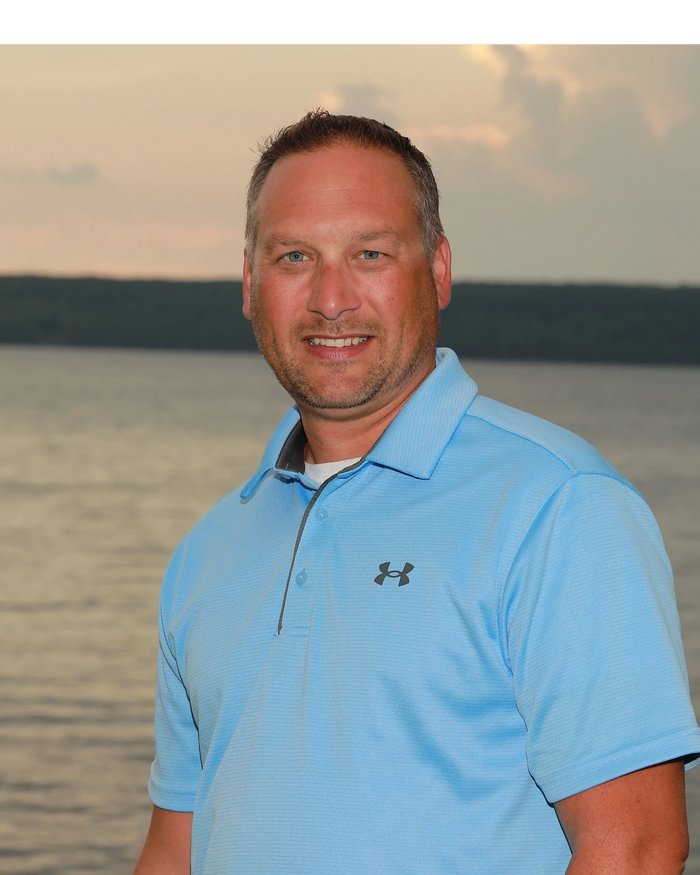 Man in a light blue Under Armour polo shirt smiling by a lake at sunset.