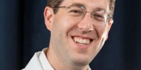 Smiling man in glasses and white coat against dark background.