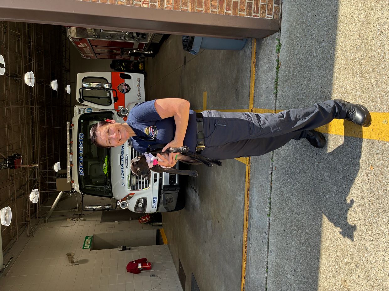 Smiling EMS worker holding a small black dog outside an ambulance in a fire station.