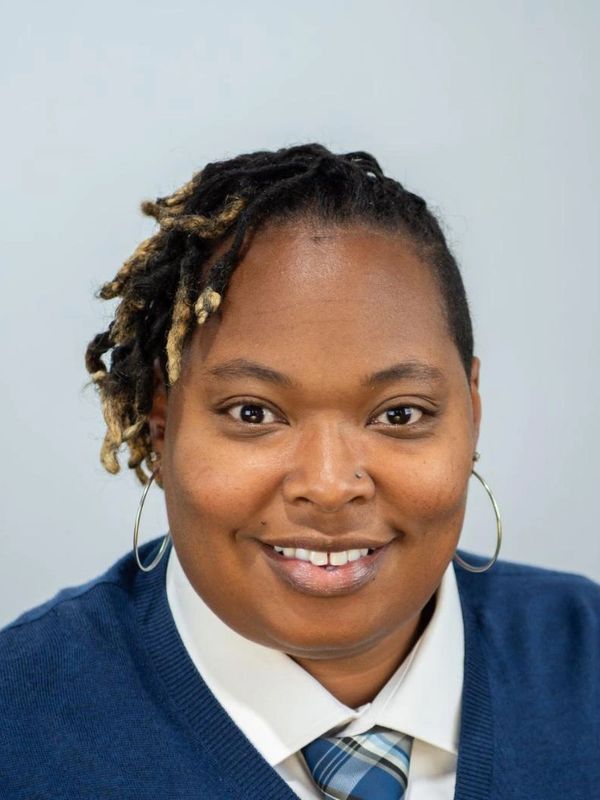 Smiling person with styled dreadlocks and hoop earrings wearing a tie and sweater.