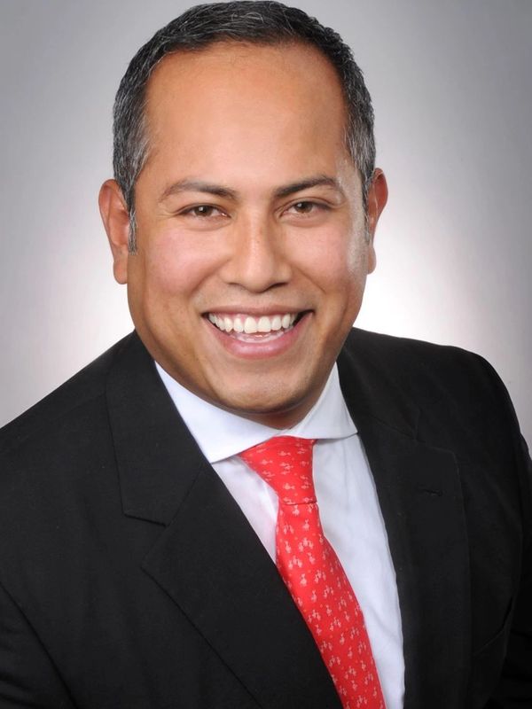 Smiling man in a black suit with a red tie against a gray background.