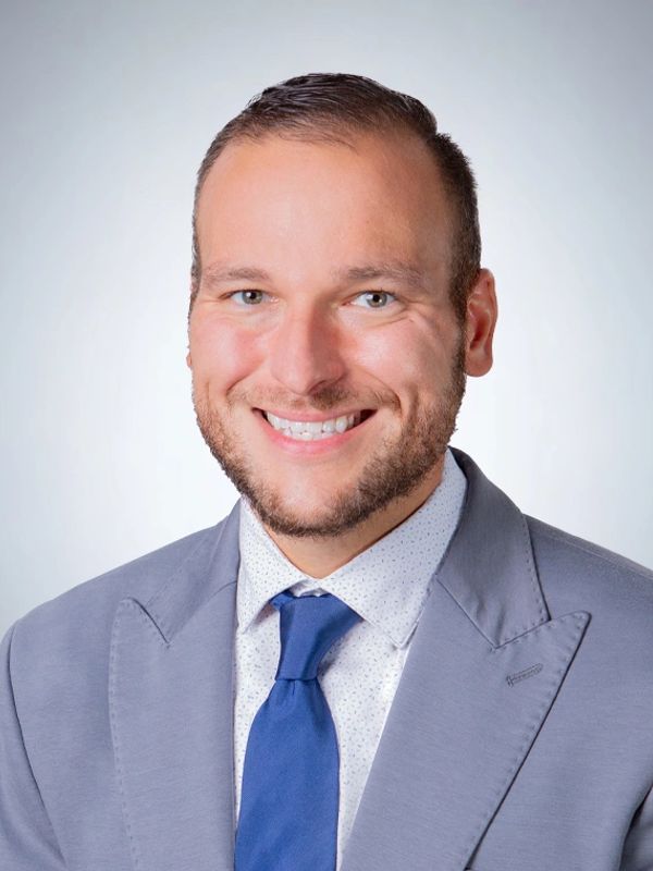Smiling man in a light gray suit and blue tie against a plain background.