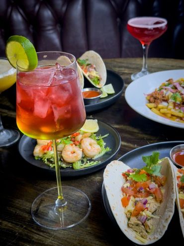 Cocktails and gourmet tacos with shrimp and fresh toppings on a wooden table.