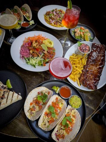 A vibrant spread of tacos, ribs, fries, and colorful drinks on a wooden table.