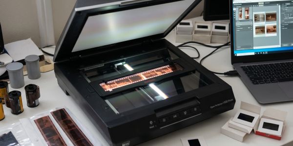 A film scanner digitizing photographic film strips on a desk with a laptop.
