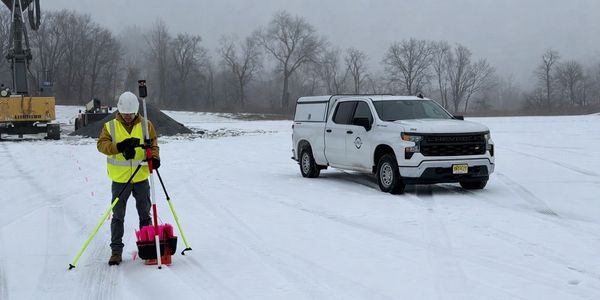 Surveyor in high-visibility gear working in snowy field next to a white pickup truck.