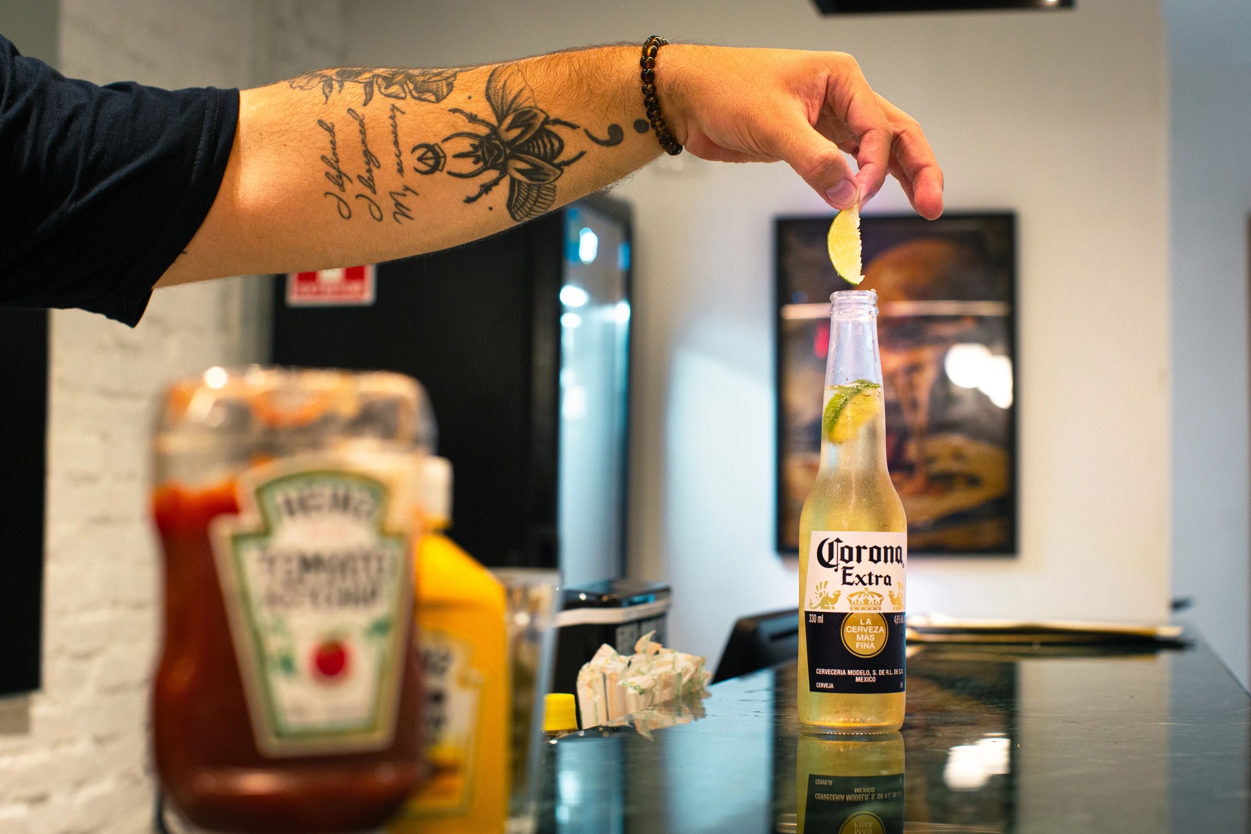 Uma garrafa de corona em cima de um balcão de um bar, com uma mão colocando uma fatia de limão