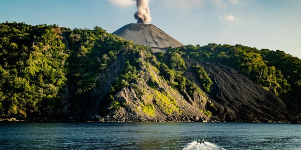 A smoking volcano behind a green forest by the water with a boat wake in the foreground.
