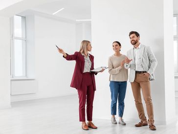 A real estate agent in a maroon suit showing a modern, unfurnished apartment to a smiling couple.