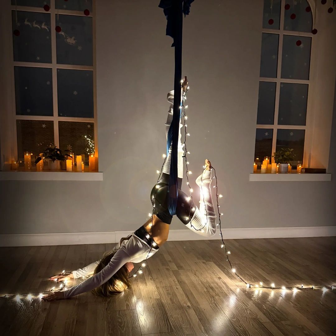 Woman performing aerial yoga upside down with string lights in a cozy room.