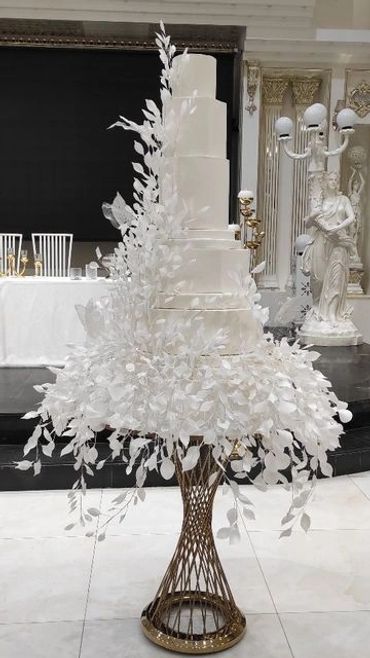 Elegant five-tier white wedding cake adorned with delicate white foliage on a gold stand.