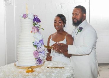Couple cutting a tall white wedding cake adorned with purple flowers.