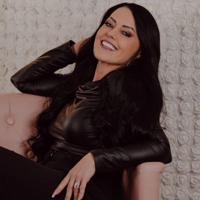 Smiling woman with long black hair in a black leather outfit, sitting on a pink chair.