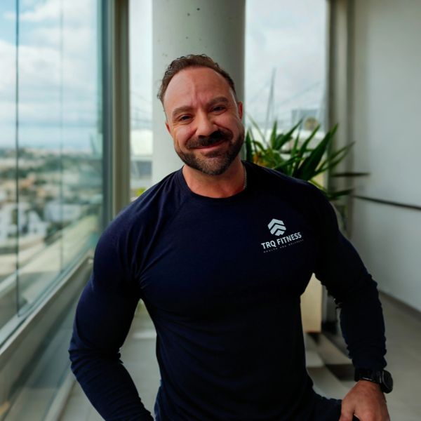 Smiling muscular man in a black TRQ Fitness shirt indoors near a window.