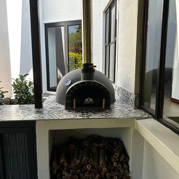 Outdoor wood-fired pizza oven with stacked firewood below and decorative tiles.
